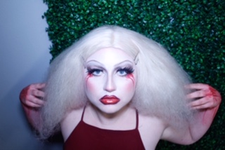 Person with dramatic white & red makeup facing camera with hands and white wig framing face.