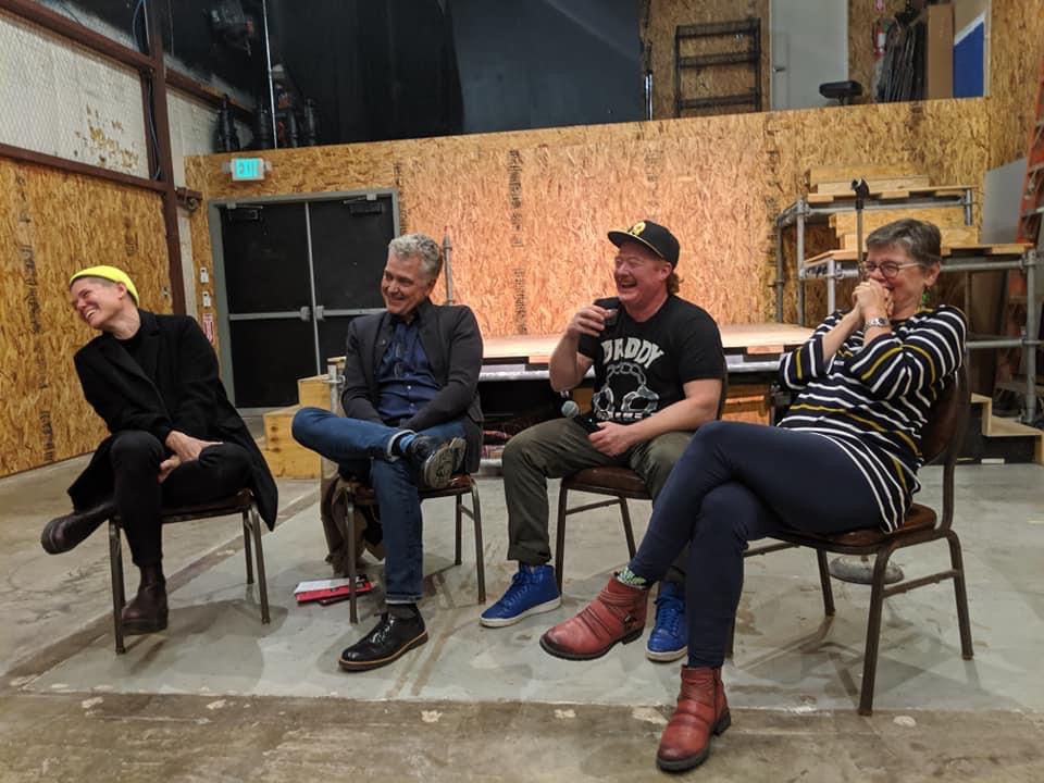 Four people siting in a semi circle in chairs in front of a stage in a theatre laughing and looking out into the audience. 