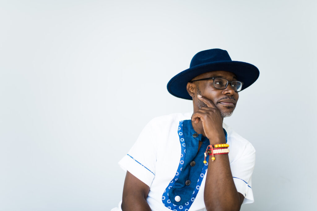 Photograph of Xavier Livermon leaning to the right, hand on chin, wearing a dark blue velvety fedora hat and a white and blue cotton short sleeved shirt