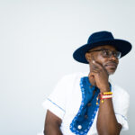 Photograph of Xavier Livermon leaning to the right, hand on chin, wearing a dark blue velvety fedora hat and a white and blue cotton short sleeved shirt