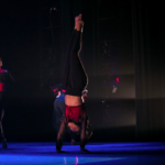 Photograph of a Black person doing a handstand on a dark stage with blue lights, wearing black pants and a black and red top. Their head is turned away from the camera.