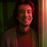 photo of author smiling at camera partly lit by pink light wearing turtle neck and cardigan