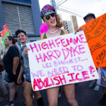 Group of people marching and holding signs. The person in the front wears a hat, necklace, and sunglasses with a sign that reads "HIGH FEMME HARD DYKE LEZBHONEST... WE NEED TO ABOLISH ICE!".