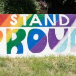 Wooden sign sitting outside with text that says "STAND PROUD". The colors behind the text represent the rainbow pride flag and the transgender pride flag.