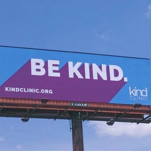 highway billboard against blue sky that says "be kind" in purple and white letters against blue background.