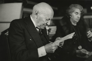 Undated photo of Isaac Bashevis Singer, with wife Alma in the background. Unidentified photographer.