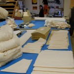 Stacks of creased board and cloth pieces ready to be sewn, a pile of completed padded cloth covers. Head of the preservation lab Apryl Voskamp, and work-study student Liz Phan consult. Photo by Mary Baughman.