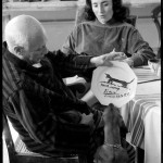 While Jacqueline Roque looks on, Lump, David Douglas Duncan's dachshund, inspects the luncheon plate Pablo Picasso has just painted for and dedicated to him. La Californie, Cannes. Gelatin silver negative. April 19th, 1957. © David Douglas Duncan.