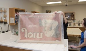 Conservators remove the poster from the bath. They are able to handle the wet paper because it is layered between strong sheets of spun polyester, called Reemay. The poster clings to the Reemay as it is pulled from the water. Photo by Pete Smith.