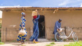 This image shows two women and a man carrying sacks of goods in front of a Nigerian structure. The women are walking, and the man has a bicycle.