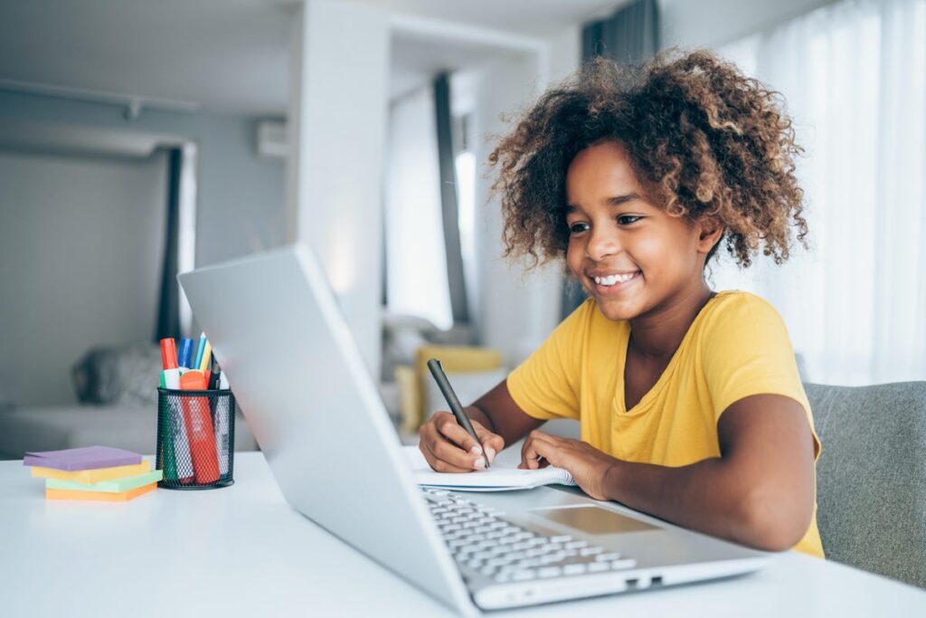 A little girl wearing a yellow shirt sitting at a desk, writing in a notebook while look at laptop screen
