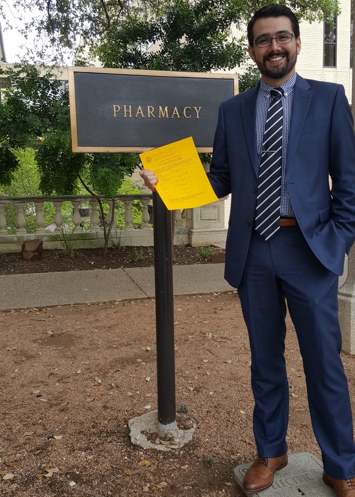 Dr. Daniel Moraga-Espinoza holding a yellow piece of paper and standing in front of the Pharmacy building sign.