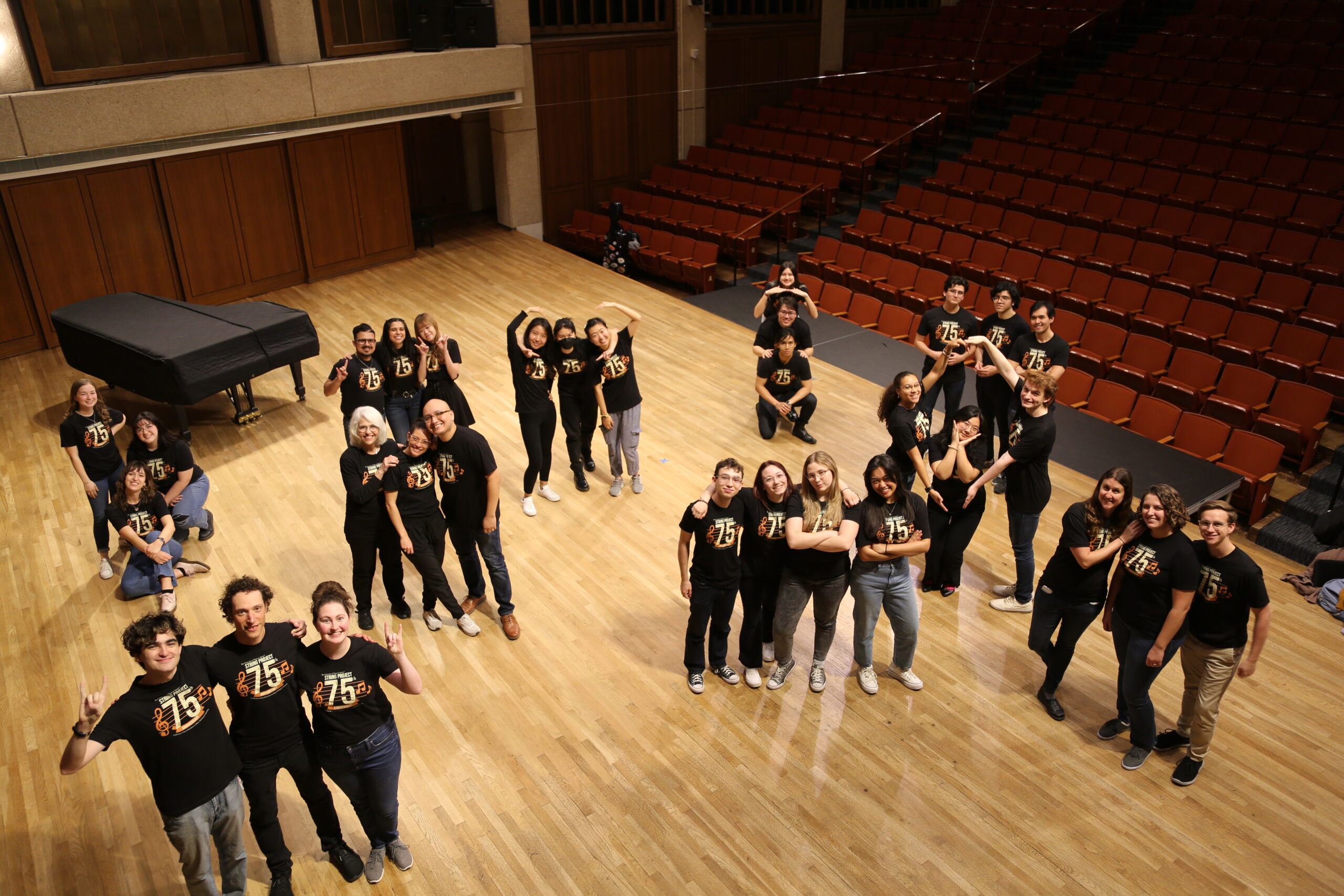 String Project students stand on stage with faculty in small groups. They're wearing 75th anniversary t-shirts.