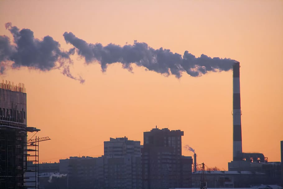 Industrial smokestacks in the midst of a cityscape, against an orange sky.