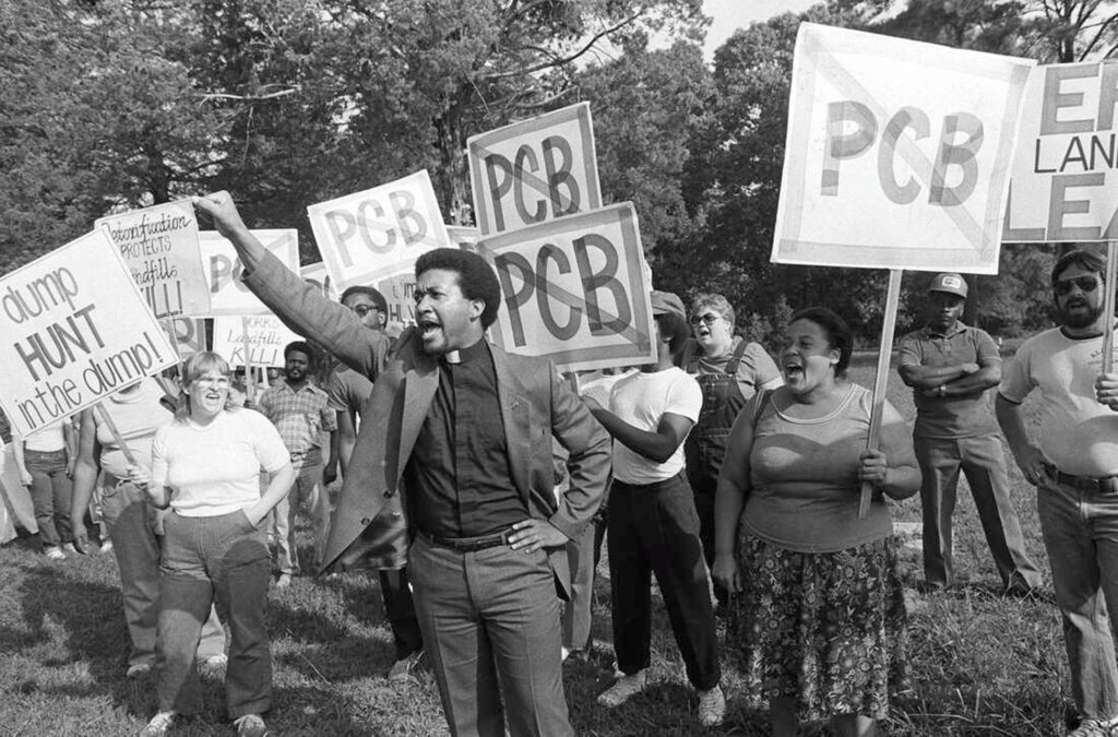Group of protesters holding "no PCB" signs in an outdoor clearing with trees behind them.