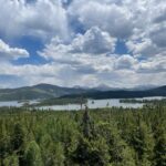Panoramic view of lake with green trees in foreground and fluffy white clouds in the blue sky.