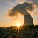 Low-angle photo of nuclear power plant emitting smoke, with sun on the horizon.