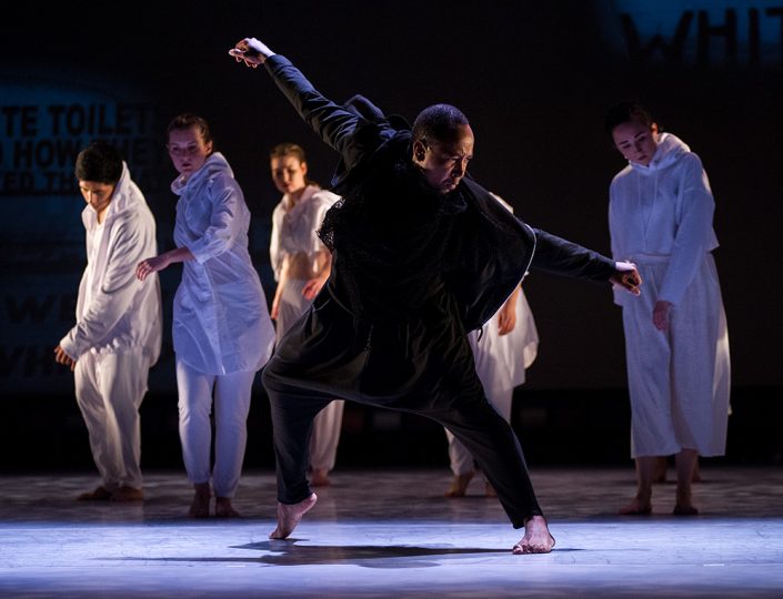 a dancer in black poses with arms outstretched with dancers in white behind him