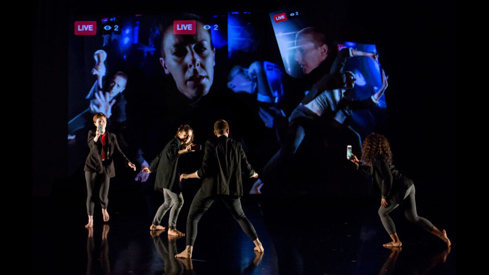 Dancer in all black poses while three other dancers record them with camera phones, with the footage they are capturing projected behind the group