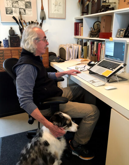man with long silver hair working at his desk with his dog sitting next to him