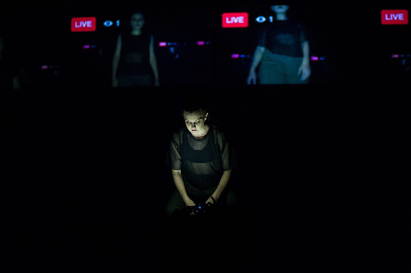 A dancer in black looks down at a phone screen, witch lights her face on the dark stage. Behind her is projected livestreams of other dancers performing.