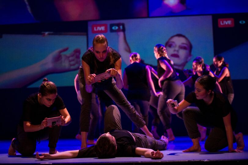 A dancer lies on the floor while three other dancers record her from different angles. In the background is another group of dancers and projections of the footage of the dancer on the floor.