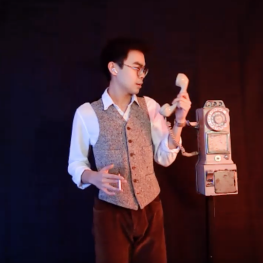 an actor with a vest and glasses on smokes a cigarette while picking up a rotary phone