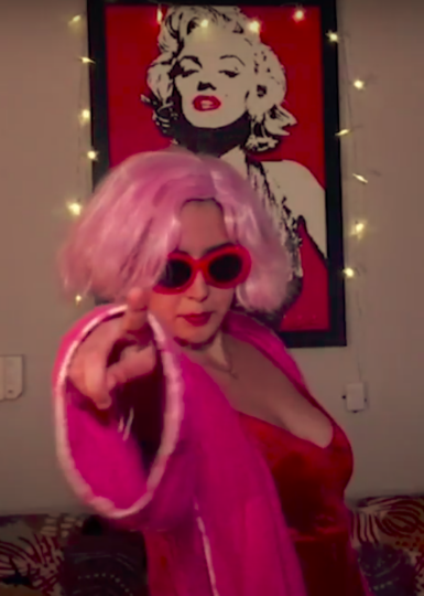 actress in a pink wig, robe and red sunglasses points at the camera