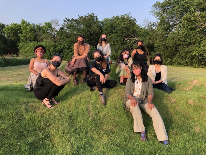 nine women wearing masks sit on a grassy hill