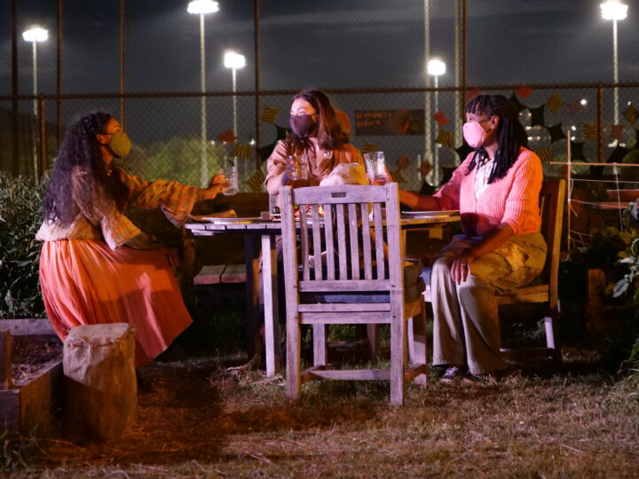 three women, all wearing masks, sit around a table in the middle of a garden