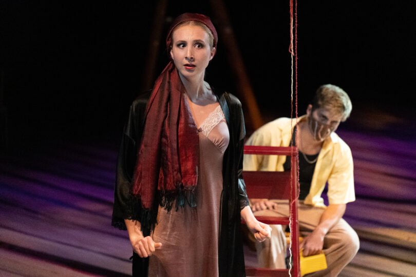 an actress with a red scarf tied around her head looks suspiciously to the side while an actor sits behind her in a chair