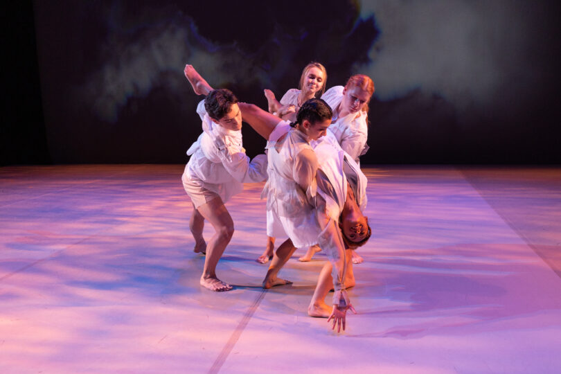 four dancers hold another dancer, who stretches her hand down to the floor