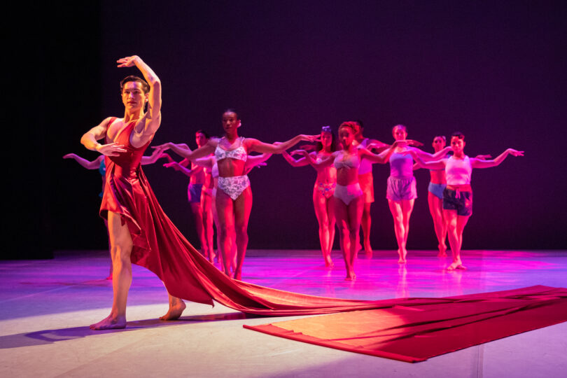dancer poses with his arms up around him and the red train of his dress laid out next to him, with an ensemble of dancers in the background