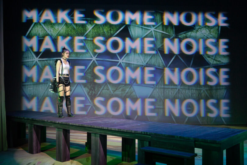 an actor in an over-the-top fighting costume stands in front of a screen that says "MAKE SOME NOISE"