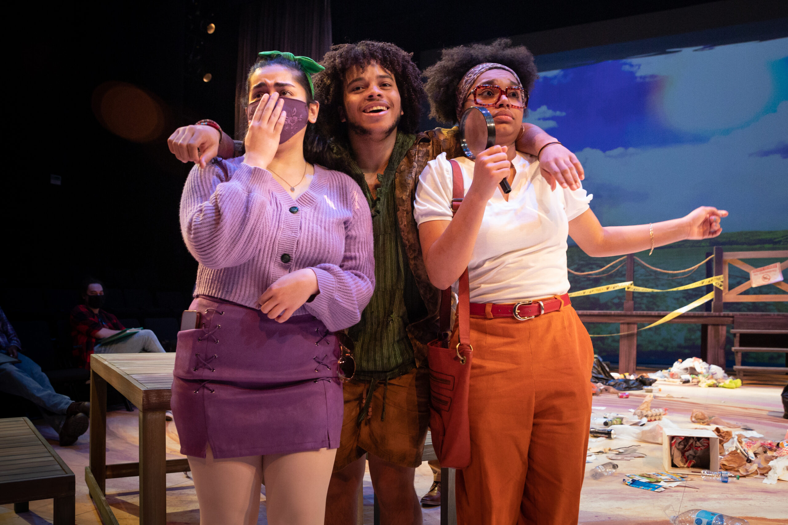 an acan actor with curly brown hair stands between two actresses, who are reacting with shocked faces