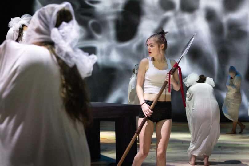 an actor gripping a double-sided spear stands in the middle of masked actresses in all white