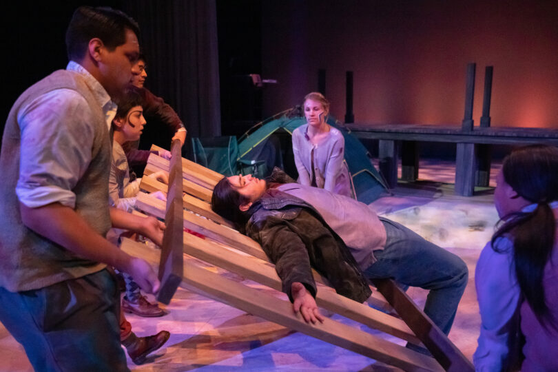 five actors hold up a structure of wooden boards, while another actor lays across it
