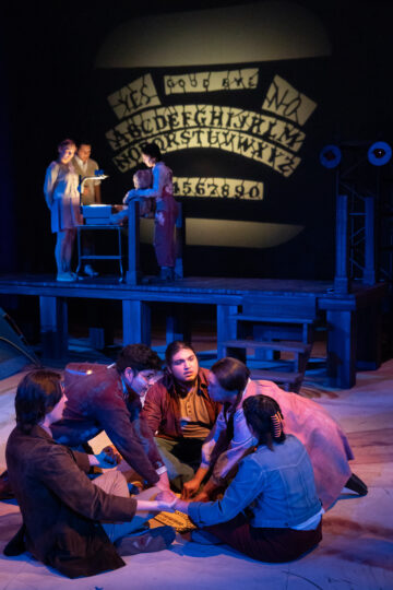 five actors sit in a circle holding hands, while three actors stand in the background manipulating a projection of an ouija board