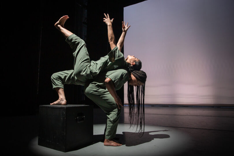 two dancers in green; one balances on a black block, leaning on the other's back and reaching up