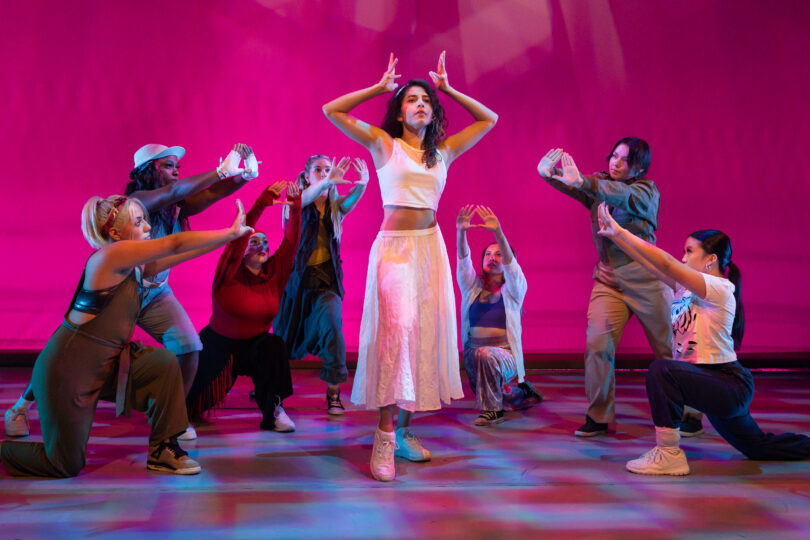 seven dancers form a semicircle around another dancers who poses with her hands above her head forming a crown