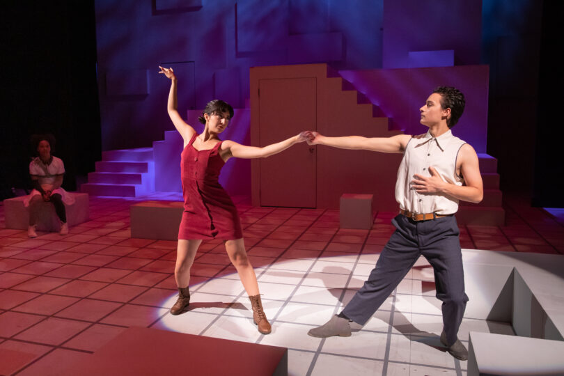 two actors dance on stage, one in a red dress and the other in a button-up without sleeves
