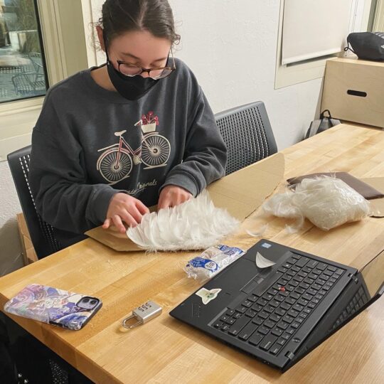 woman fabricating fake wings by gluing white feathers onto a piece of cardboard