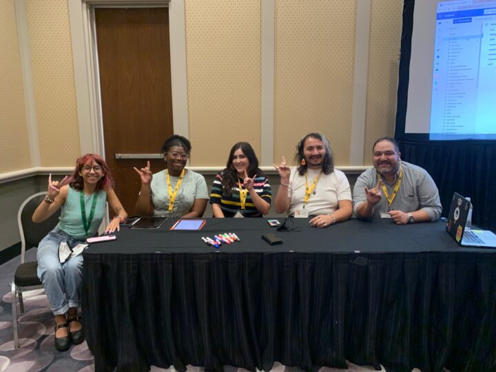 four students and faculty member Michael Ávila sit behind a table, posing while making the "Hook 'em horns" sign