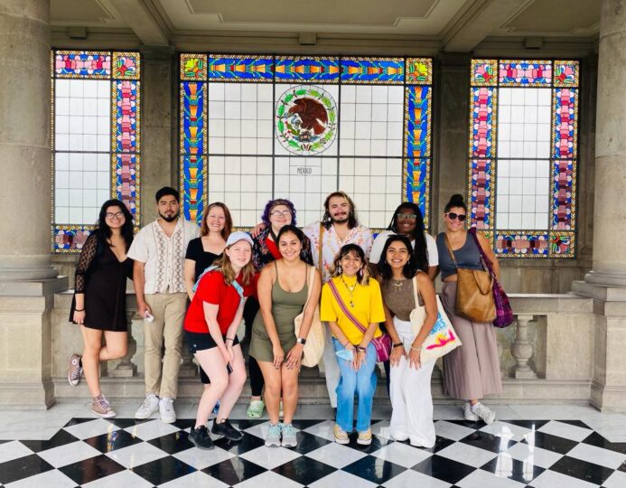 nine students pose with Roxanne Schroeder-Arce and Leslie Moody Castro in front of a decorative window featuring the Mexican coat of arms