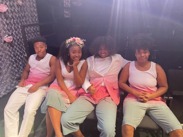 four actors in dyed pink, white and green costumes sit in the audience together