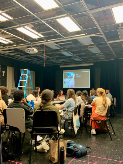 About 20 people sit around a ring of tables, bags, and chairs scattered around. Their attention is directed toward a projection screen that features an image of women in pink jumpsuits. 