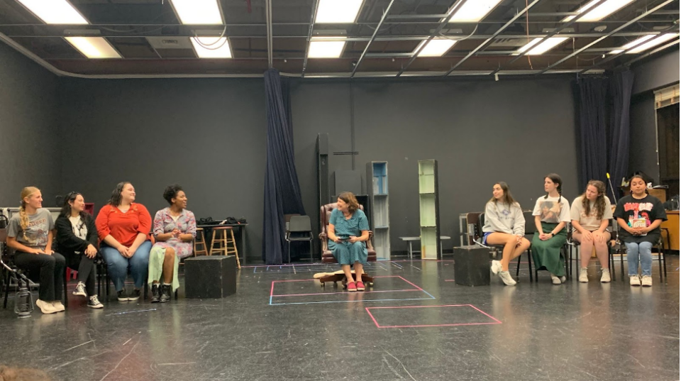 Two sets of four actors sit on either side of Barbara. The group is in a dark rehearsal room lit with fluorescent lights. Colorful tape covers the floor. 