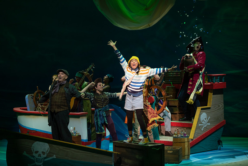 A pirate crew sings on a ship with Finley, illuminated and standing tall