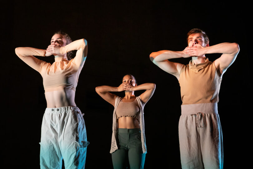 Three ballet dancers wearing minimalist earth toned apparel stand with their hands covering their mouths. 