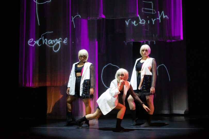 Three dancers wearing white blazers, leather skirts and white bob wigs pose dramatically